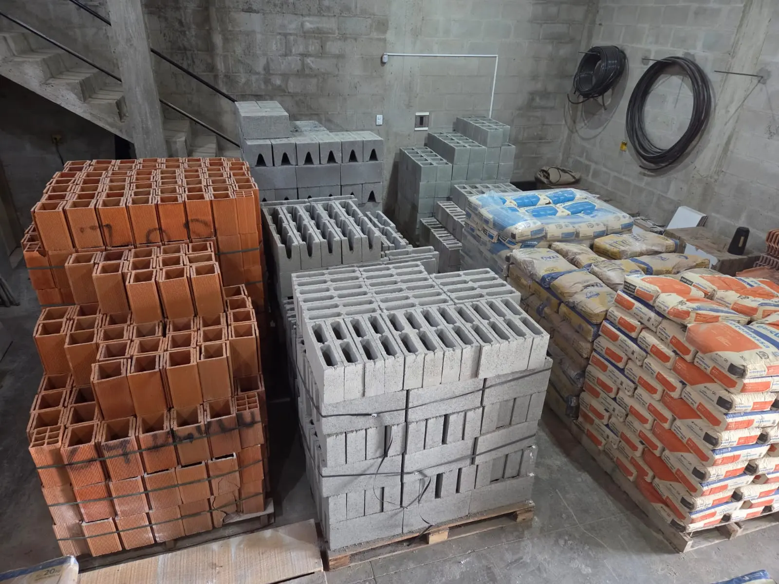 Tijolos cerâmicos, blocos de concreto e sacos de cimento empilhados em um depósito, com rolos de fio elétrico ao fundo.
