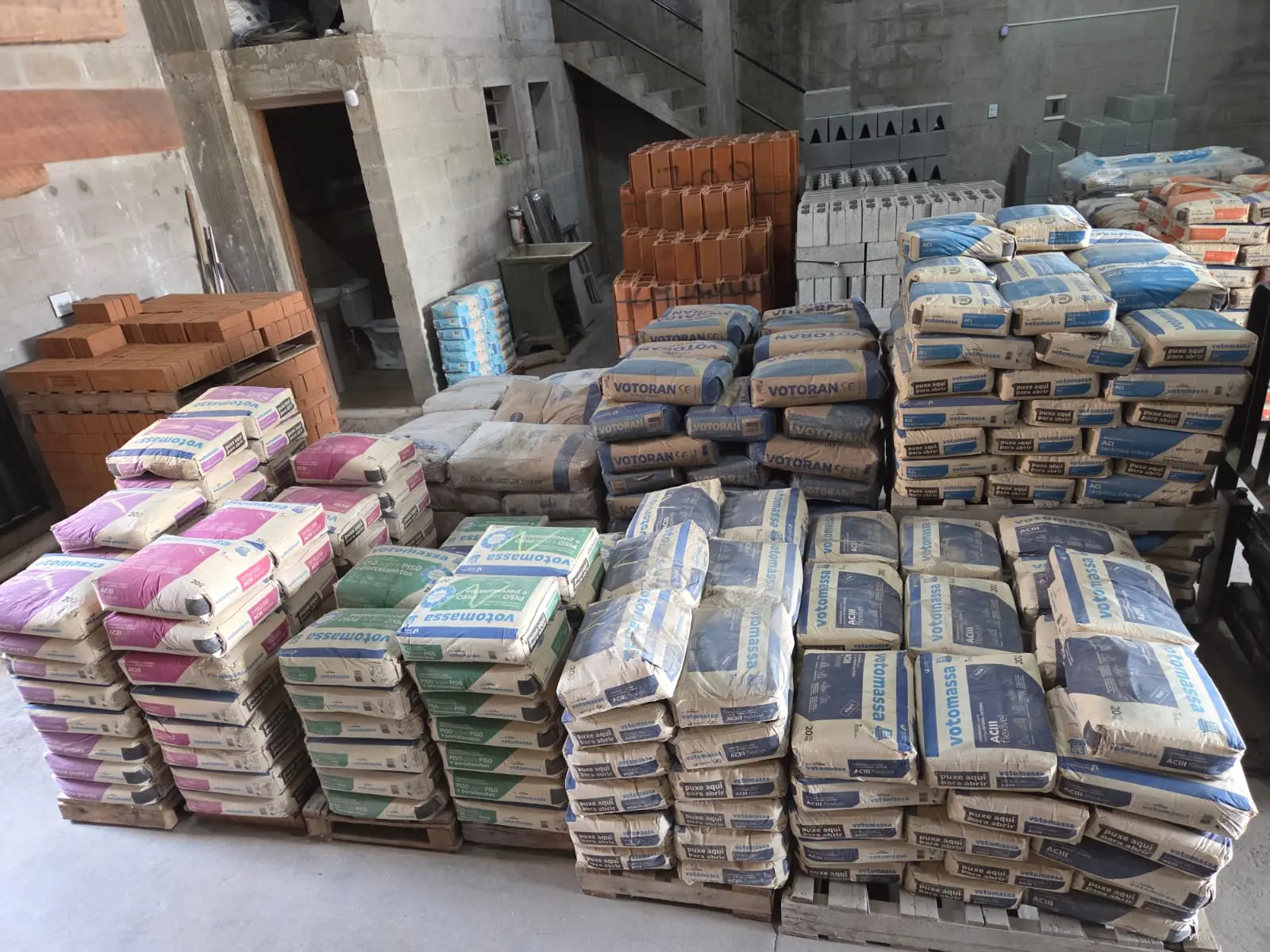 Pallets com sacos de cimento Votoran e argamassa Votomassa em diferentes cores, ao lado de tijolos e blocos de concreto empilhados.
